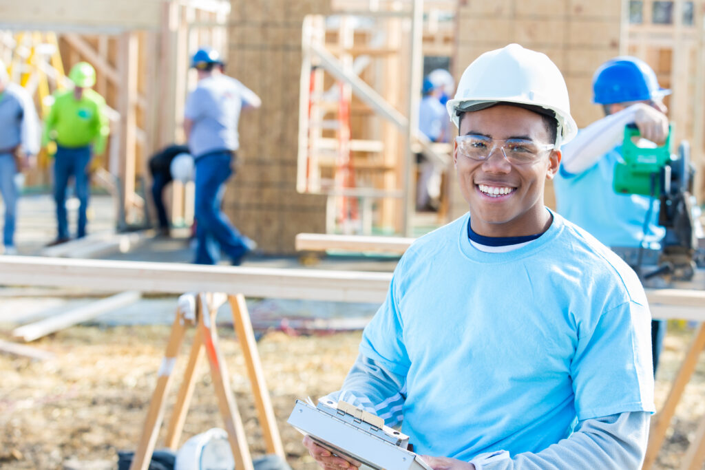 Young African American male construction foreman smiles confidently at new home construction site. He is wearing a white hard hatand safety glasses. Construction workers are working in the background.